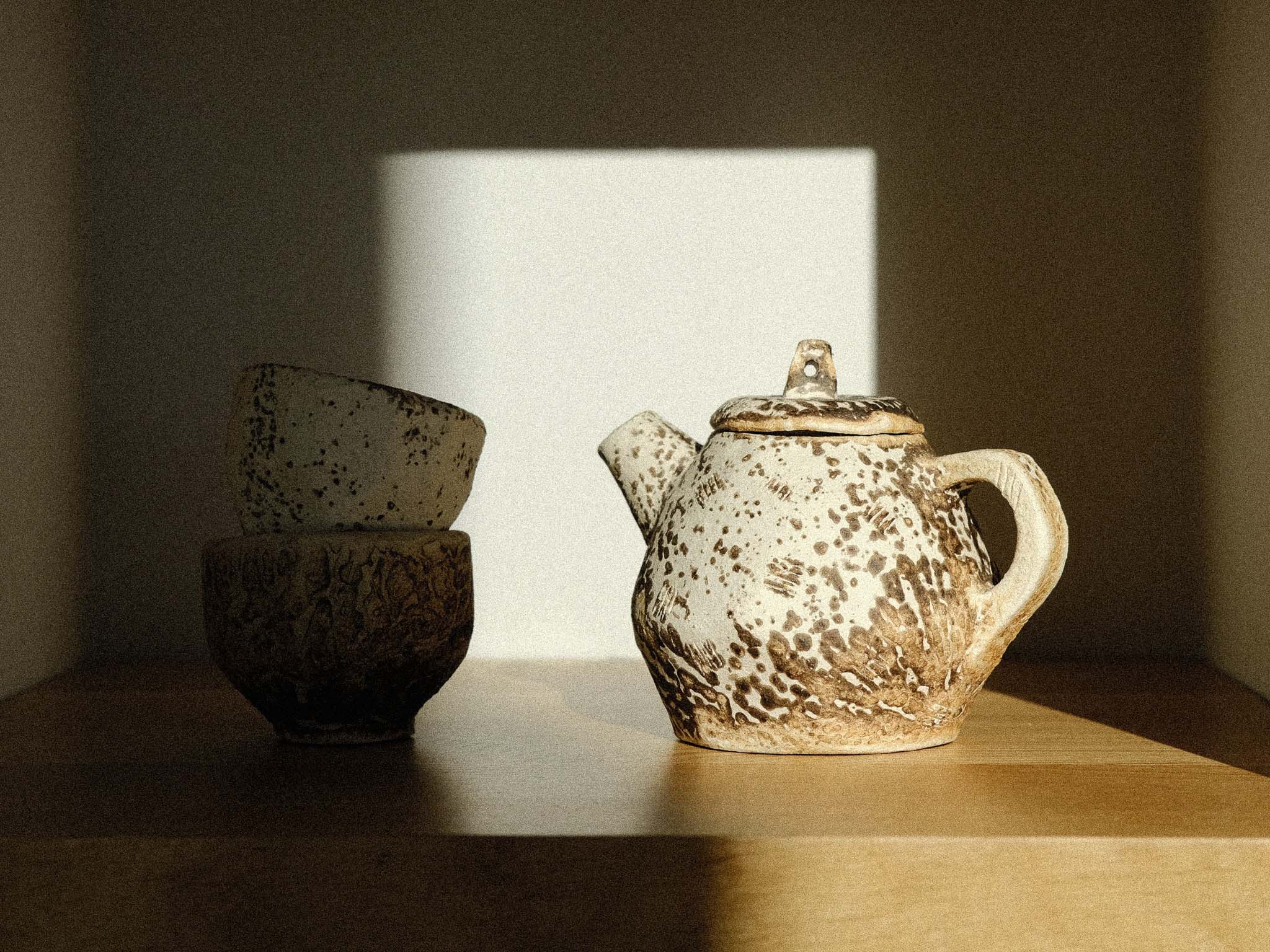 Elegant kurinuki cups and small teapot made with Baltic raku technique on wooden shelf in modern white interior, bathed in soft sunlight and shadow play.