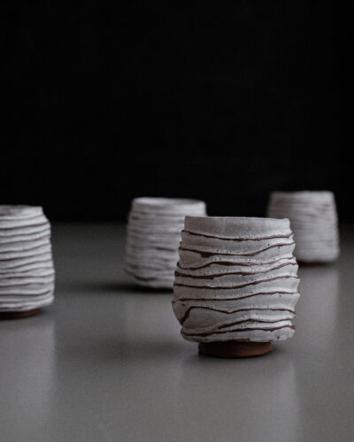 Kurinuki Cup Nº5 with white and iron red-brown decor, carved to evoke dunes or pine cones, set against a dark background, showcasing its textured, wabi-sabi imperfections.