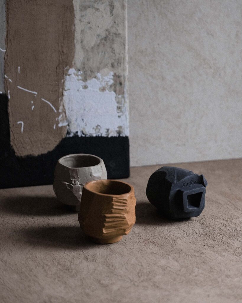 Ondou studio slowmade ceramics "Unfired Kurinuki Cup Nº1, Nº2 and Nº4 surrounded by clay paints and a backdrop painted with clay, creating a tranquil, warm atmosphere that highlights the raw beauty of the piece.