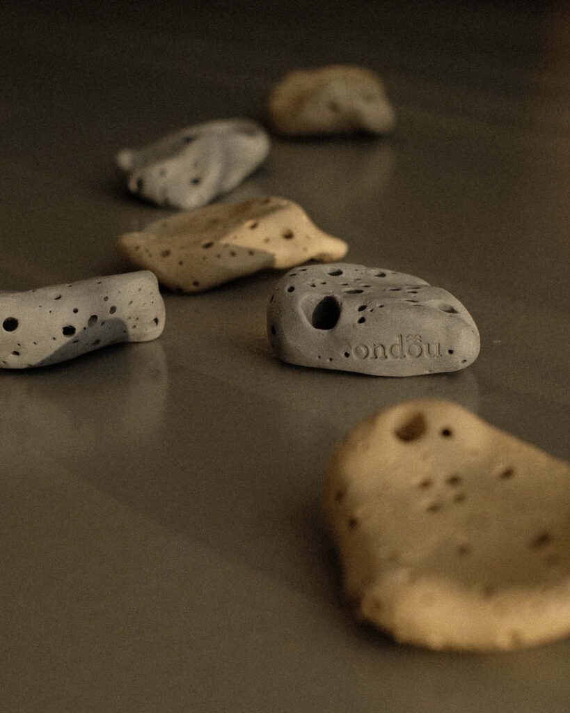 Ondou studio slowmade ceramics Sculptural incense pebble burners in dark and beige tones scattered on grey concrete, with sunlight creating soft, organic shadows.
