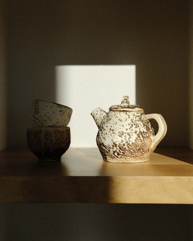 Ondou studio slowmade ceramics Elegant kurinuki cups and small teapot made with Baltic raku technique on wooden shelf in modern white interior, bathed in soft sunlight and shadow play.