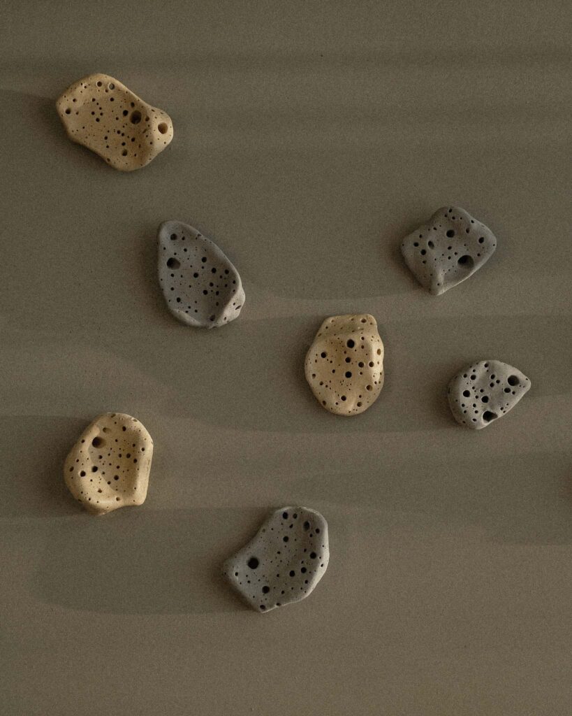 Ondou studio slowmade ceramics Sculptural incense pebble burners in dark and beige tones scattered on grey concrete, with sunlight creating soft, organic shadows.