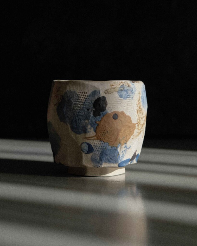 Light beige kurinuki cups with abstract expressionist painting in beige, brown, blue, and black tones, incorporating wild clay and sand for texture, captured in strong light against a dark background.