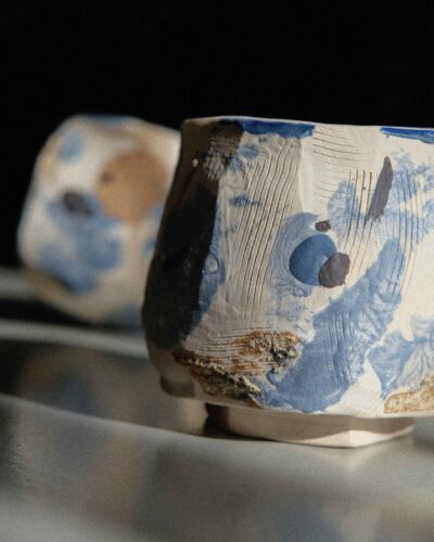 Light beige kurinuki cups with abstract expressionist painting in beige, brown, blue, and black tones, incorporating wild clay and sand for texture, captured in strong light against a dark background.