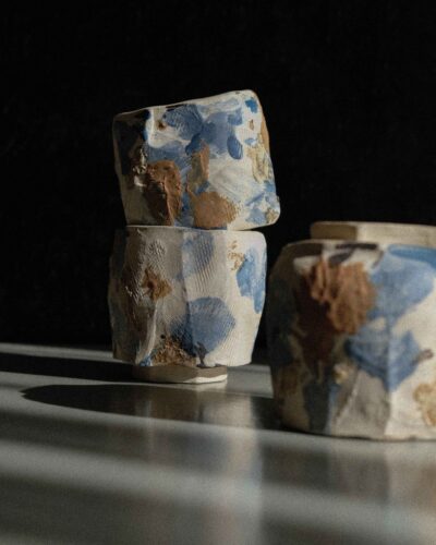 Light beige kurinuki cups with abstract expressionist painting in beige, brown, blue, and black tones, incorporating wild clay and sand for texture, captured in strong light against a dark background.