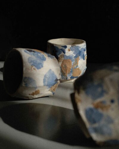 Light beige kurinuki cups with abstract expressionist painting in beige, brown, blue, and black tones, incorporating wild clay and sand for texture, captured in strong light against a dark background.