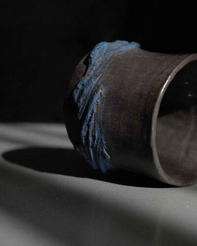 Rankų darbo juodas keramikinis puodelis, pagamintas kurinuki technika, su iškiliais mėlynais išpjaustymais, nufotografuotas dramatiškoje šviesoje.