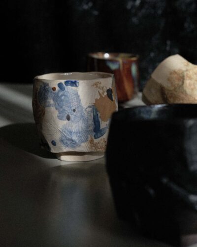 Light beige kurinuki cups with abstract expressionist painting in beige, brown, blue, and black tones, incorporating wild clay and sand for texture, captured in strong light against a dark background.