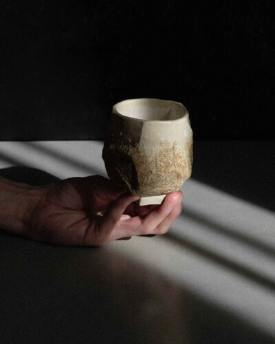 Sculptural Ondou kurinuki cup no.12 from light beige clay, strongly carved and decorated with local wild clay and sand mixture. Photographed in a dramatic light against the dark background.