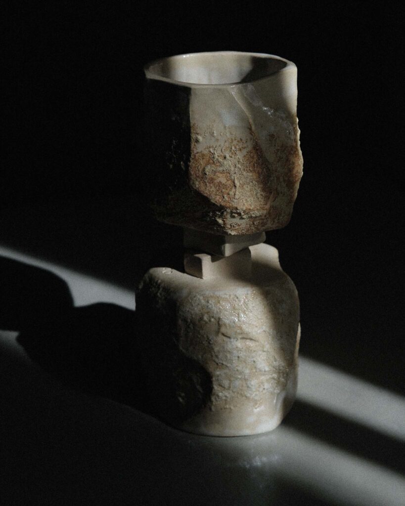 Two geometrically carved kurinuki cups from light beige wild clay stacked foot-to-foot, forming a sculptural composition with star-like bases, photographed in strong light against a dark background for dramatic contrast. Du geometriškai išpjaustyti kurinuki rankų darbo puodeliai iš šviesios smėlio spalvos akmens masės molio, formuojantys skulptūrinę kompoziciją, nufotografuoti stiprioje šviesoje tamsiame fone, sukuriant dramatišką kontrastą.