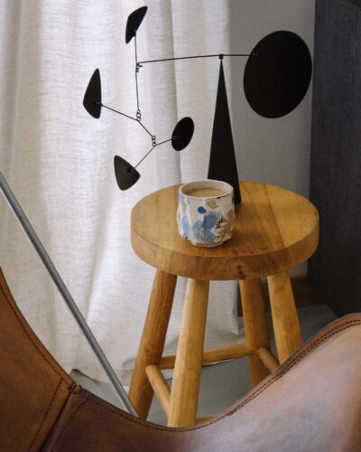 Kurinuki Cup Nº11 on a teak wood stool in a minimal wabi-sabi interior with a black mobile sculpture, vintage leather butterfly chair, and linen curtain in soft light.