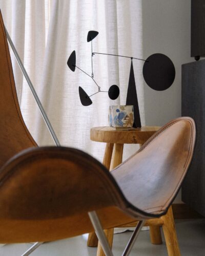 Kurinuki Cup Nº11 on a teak wood stool in a minimal wabi-sabi interior with a black mobile sculpture, vintage leather butterfly chair, and linen curtain in soft light.