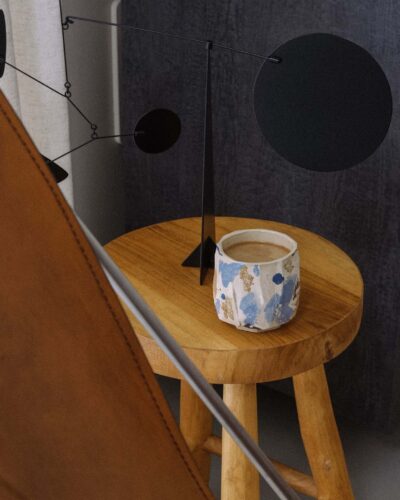 Kurinuki Cup Nº11 on a teak wood stool in a minimal wabi-sabi interior with a black mobile sculpture, vintage leather butterfly chair, and linen curtain in soft light.