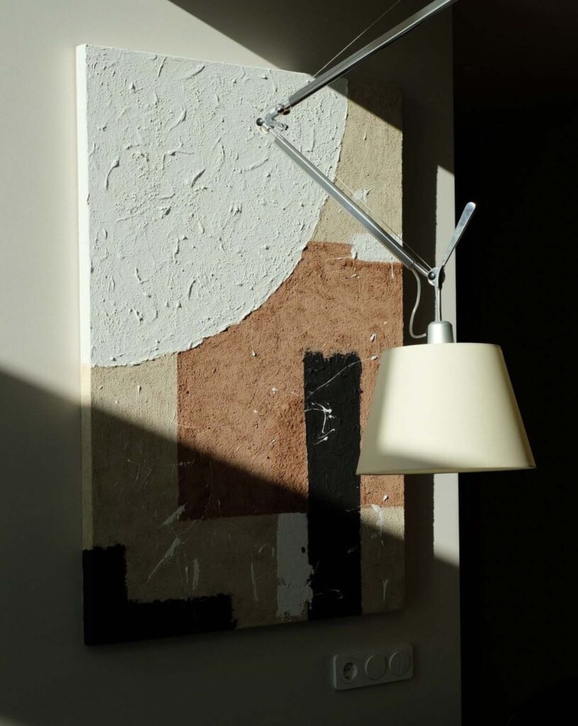 Ondou studio slowmade ceramics Textural abstract clay painting in neutral tones of white, brown, beige, and black hanging in a minimal interior, partially lit by sunlight and Artemide ceiling lamp.
